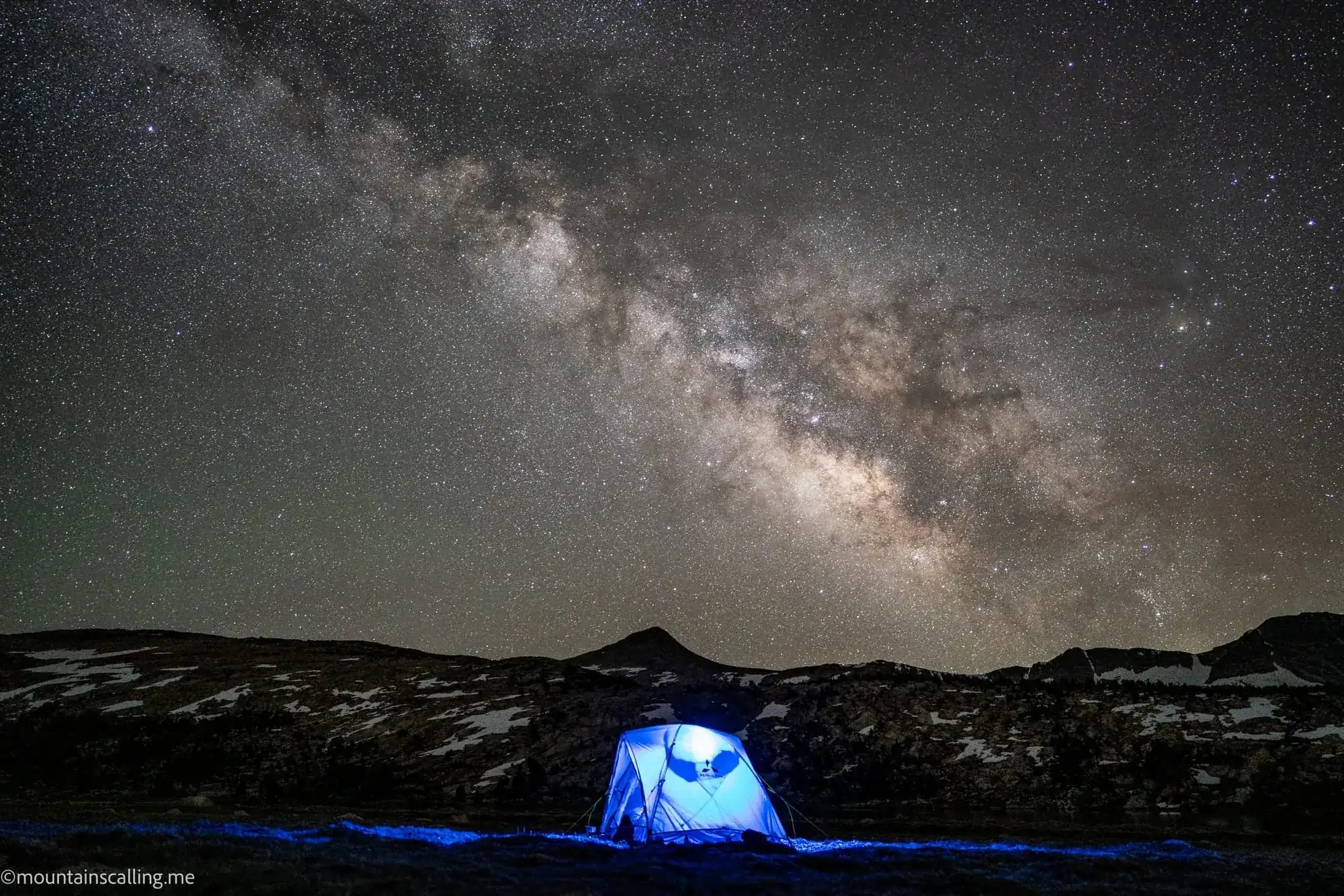 Milky Way galaxy arcing over illuminated tent at backcountry campsite in Yosemite high country with mountain silhouettes