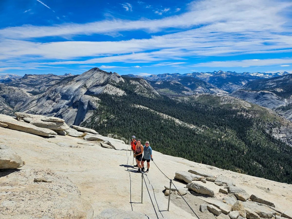 Three hikers celebrating on Half Dome's summit with cables and Yosemite's granite peaks stretching to the horizon