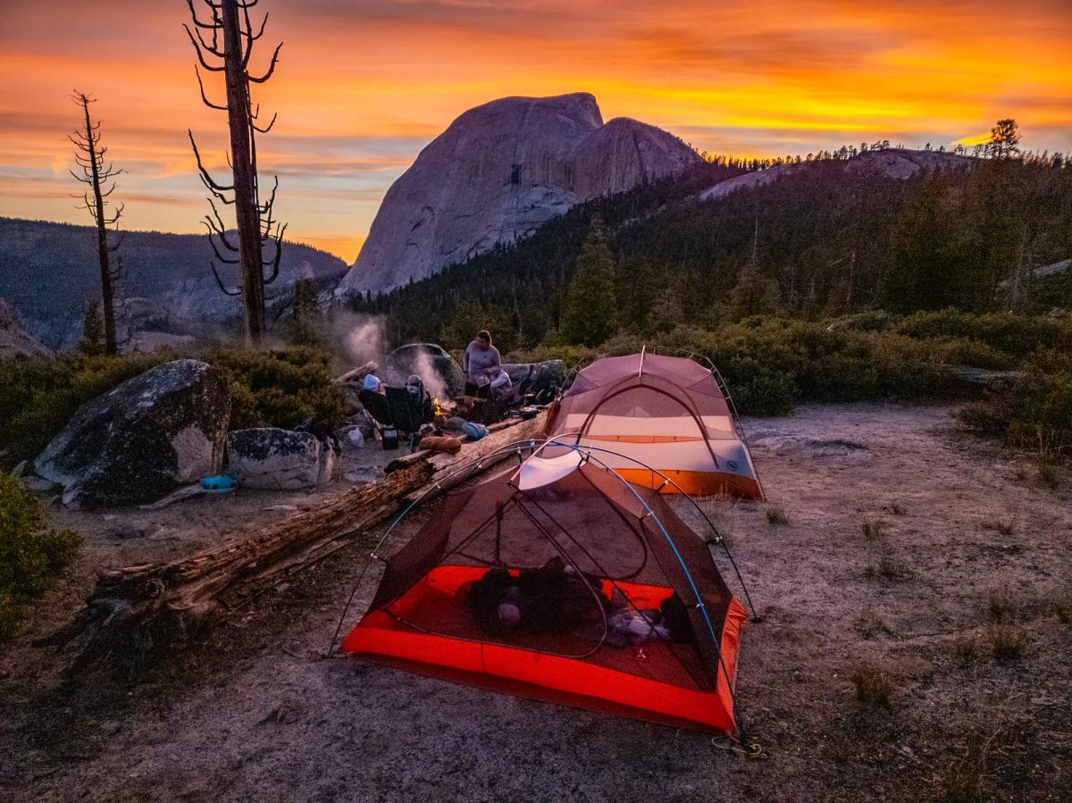 Professional Yosemite guide leading backpacking trip with illuminated tent and campfire at sunset with Half Dome