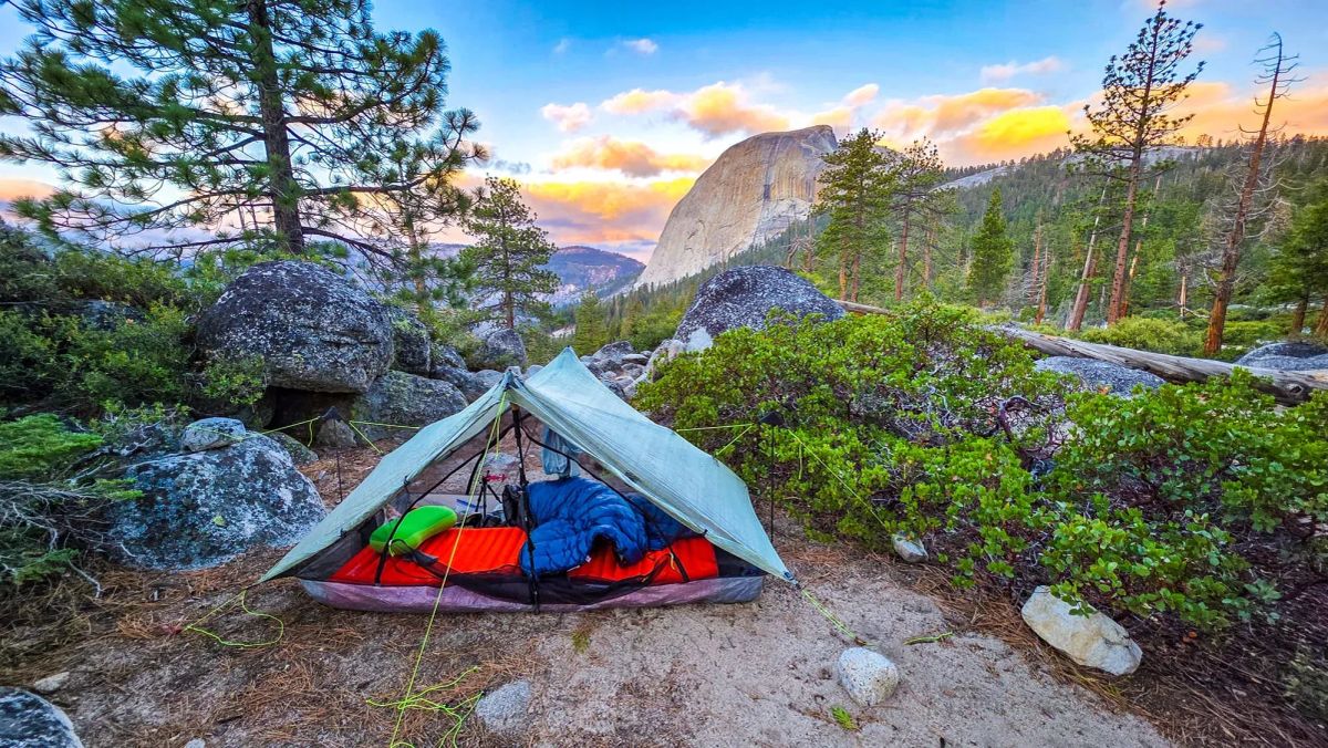Backpacking camp with tarp shelter and sleeping bags in Yosemite wilderness with Half Dome visible at sunset