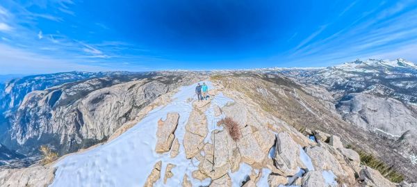 Hikers standing on the snow-patched granite summit of Clouds Rest in Yosemite at 9,926 feet with panoramic Sierra Nevada views and dramatic blue sky with wispy clouds
