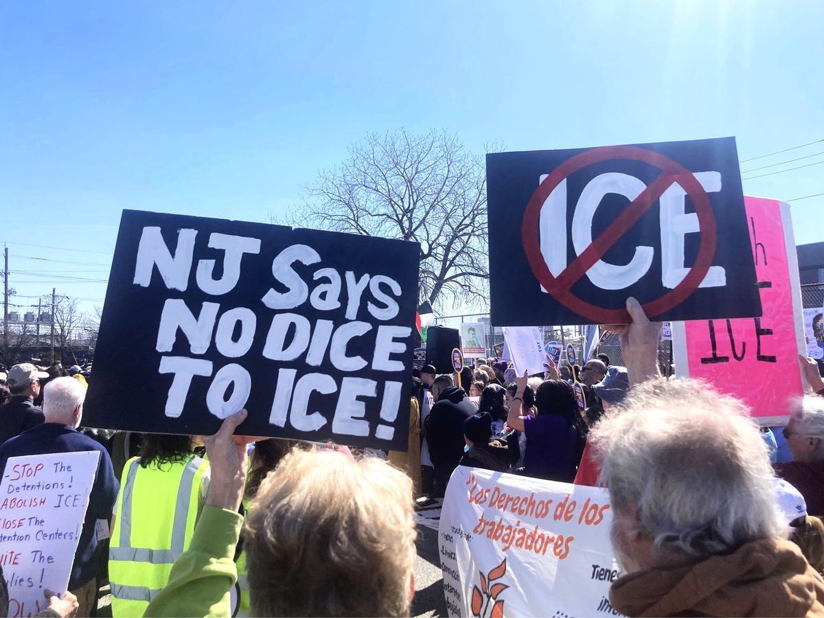 Under a blue sky, protesters hold up signs with anti-ICE logos, such as NJ SAYS NO DICE TO ICE!