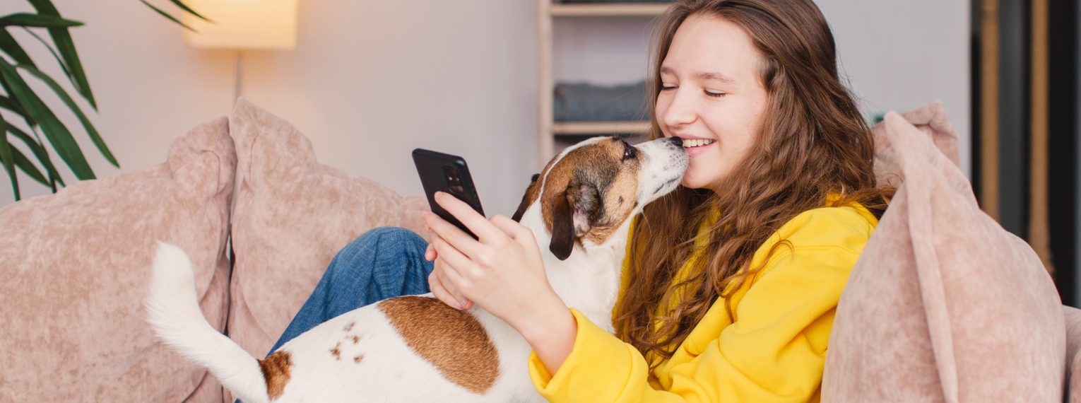 Eine Hundebesitzerin schmust mit ihrem Hund auf dem Sofa und zahlt währenddessen auf dem Handy eine Tierarztrechnung.