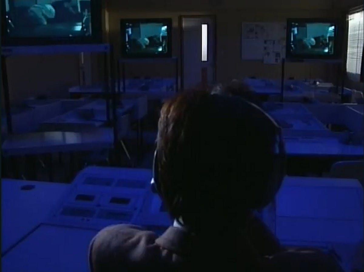 fujimura, from behind, watches a sexual assault play out on three separate televisions in a dark classroom