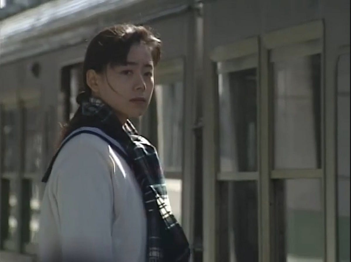 mayu, who is wearing a scarf, stands in front of a train