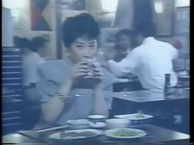 A woman drinks from a cup. In front of her are various dishes of food. She sits in a restaurant; various customers can be seen sitting at tables behind her.
