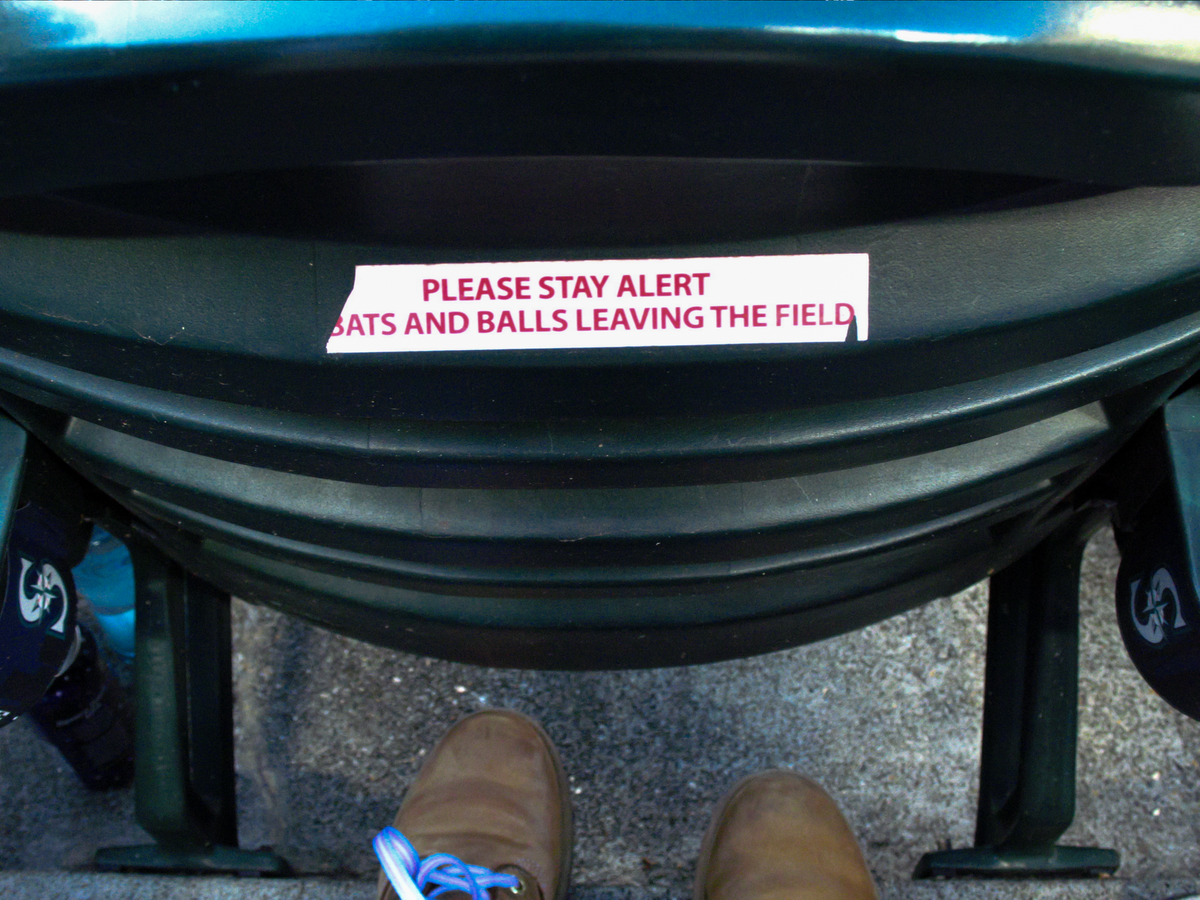 The back of a seat in a baseball stadium, with a white sticker with red text reading "PLEASE STAY ALERT BATS AND BALLS LEAVING THE FIELD".