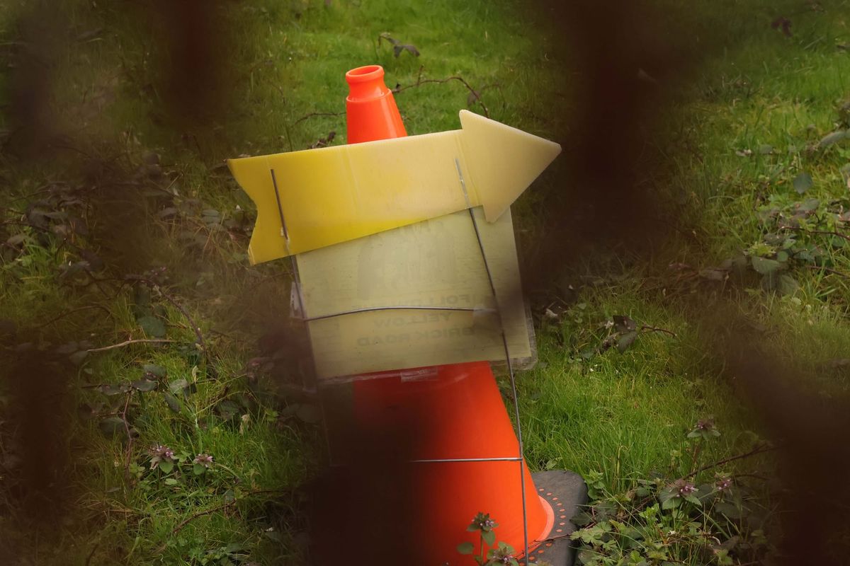 A yellow arrow sign pointing right in front of an orange traffic cone, photographed through a rusty chain-link fence.
