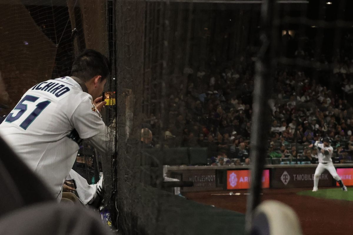 A fan in an Ichiro jersey photographs the batting area with their phone. Out of focus, a Mariners batter can be seen warming up.