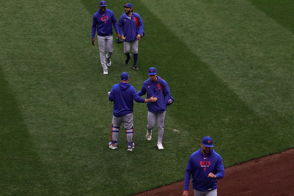 Four pitchers in Mets gear cross the grass of T-Mobile park, walking towards the warning track. One high-fives the bullpen catcher, who is facing away from the camera.