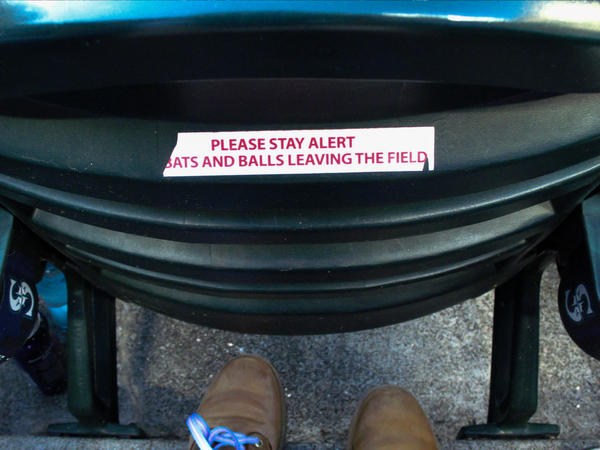 The back of a seat in a baseball stadium, with a white sticker with red text reading "PLEASE STAY ALERT BATS AND BALLS LEAVING THE FIELD".