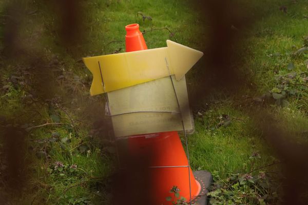 A yellow arrow sign pointing right in front of an orange traffic cone, photographed through a rusty chain-link fence.