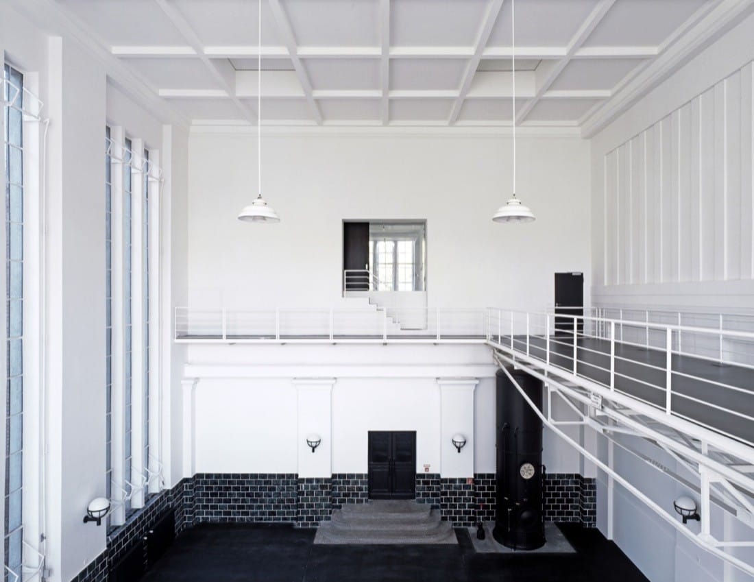 Main hall with white walls, black floor, mezzanine railings and tall industrial windows.
