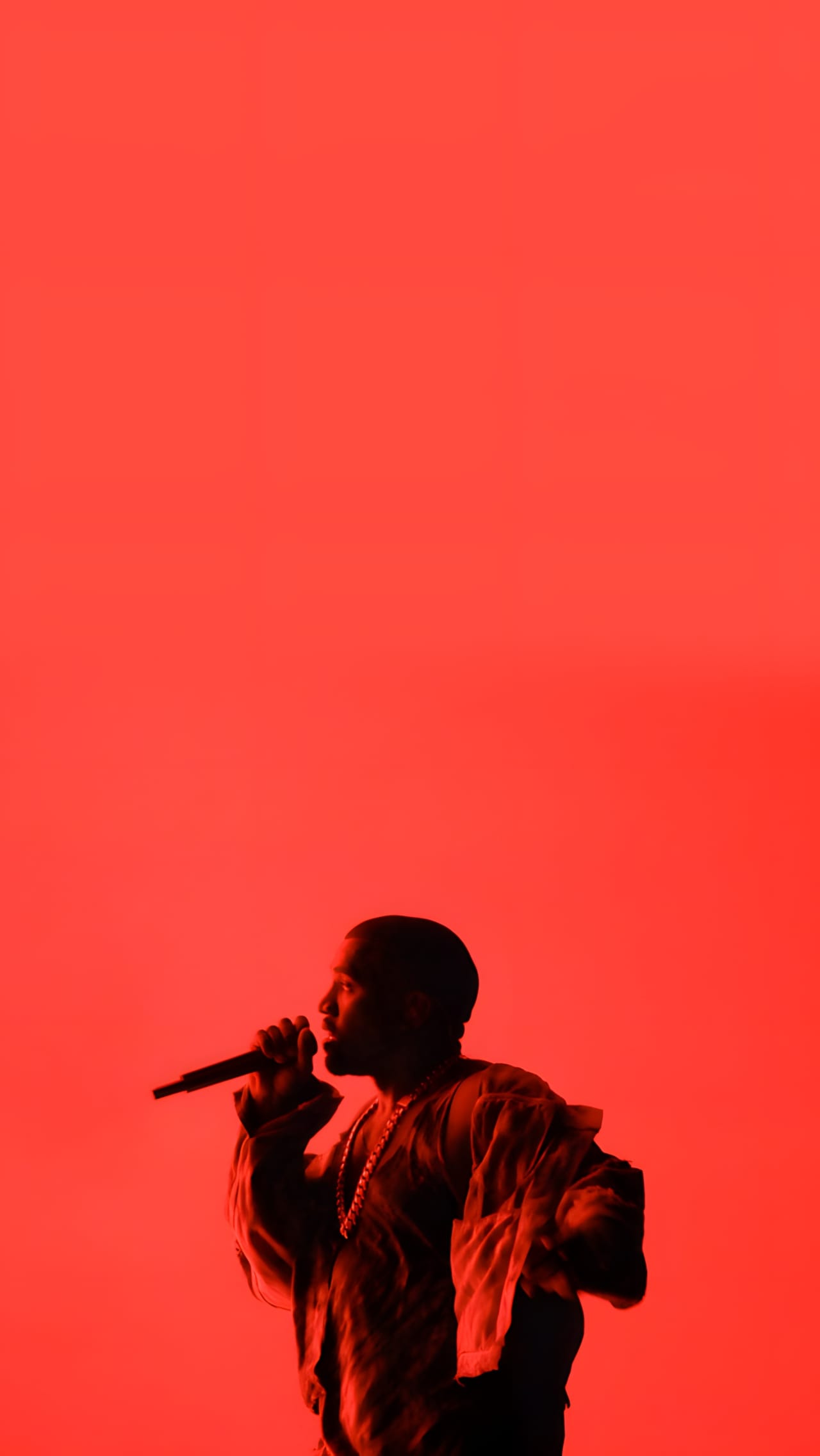 Kanye West performing on the Yeezus Tour in red stage lighting, singing into a microphone while wearing a layered Maison Martin Margiela coat.