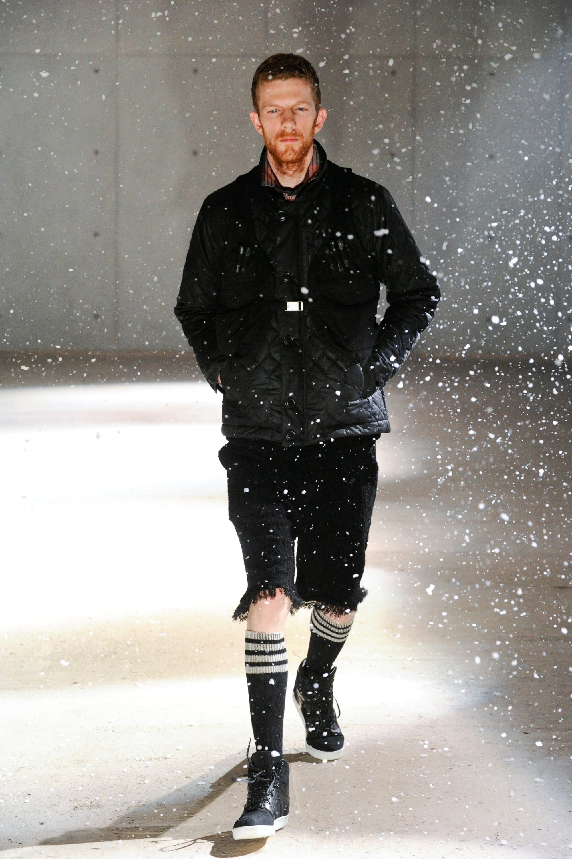 White Mountaineering Wilderness Outfitters look 2: black quilted field jacket, tactical vest, cropped trousers and striped socks in artificial snow.