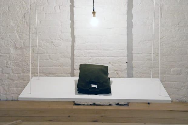 Folded olive Silent Ventures knitwear displayed on a suspended white shelf beneath a bare bulb.