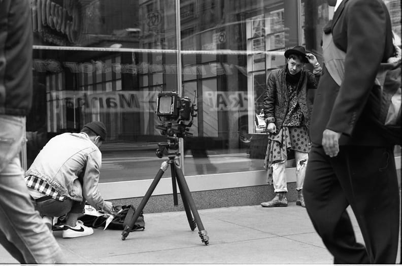 Camera crew photographing a model in a hat and layered patterned garments beside a shop window.