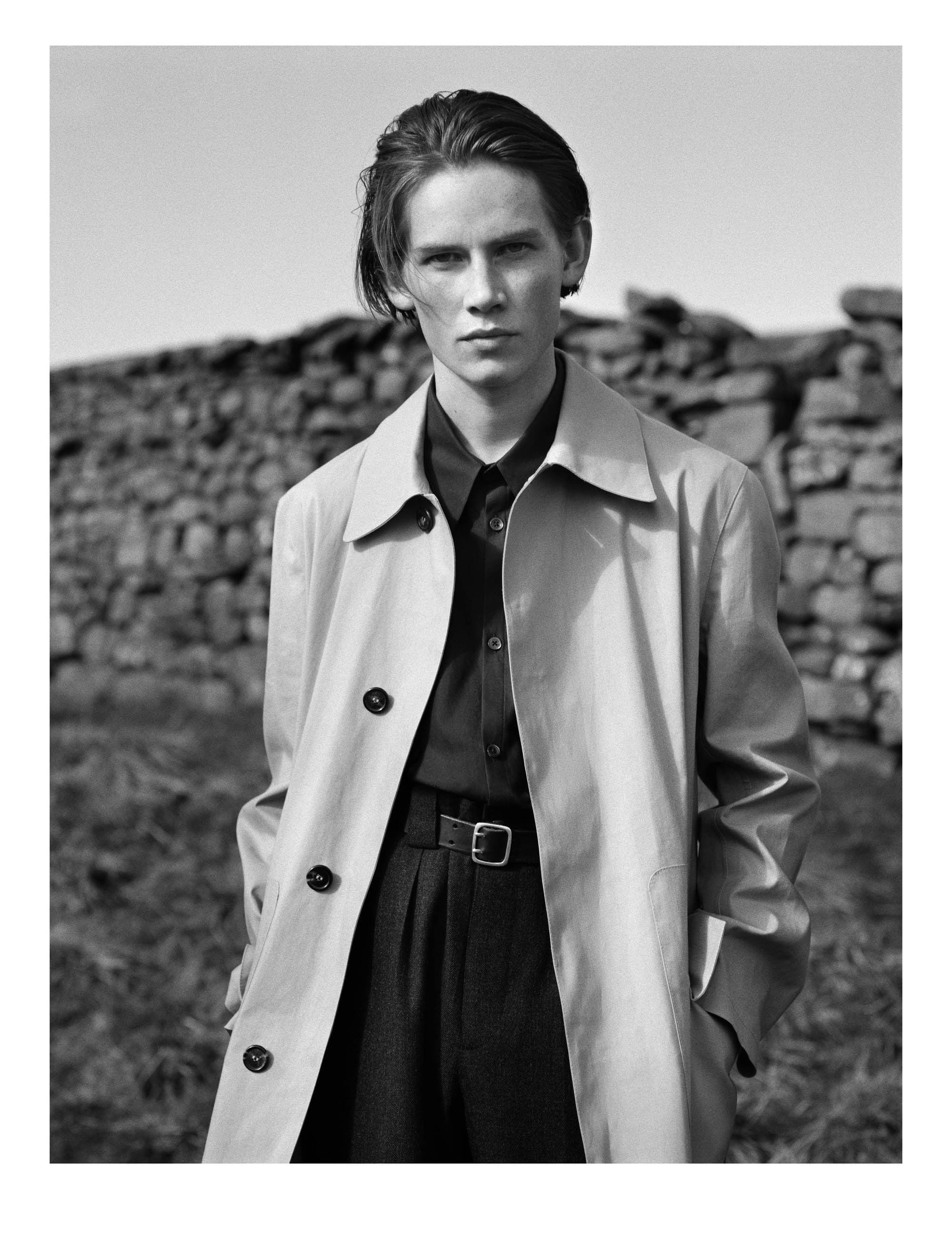 Gustav Frich wearing a light Margaret Howell raincoat over dark tailoring beside a dry-stone wall.