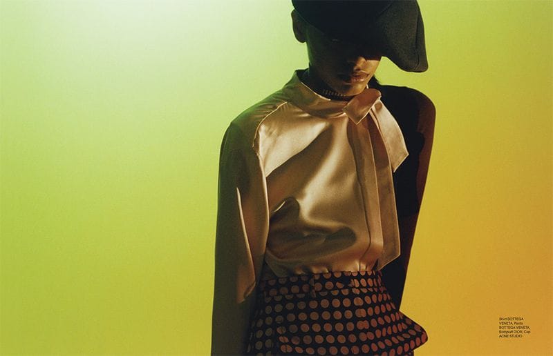 108 Big Optics spread with a model in a wide black hat, pale satin blouse and patterned skirt against green and yellow light.