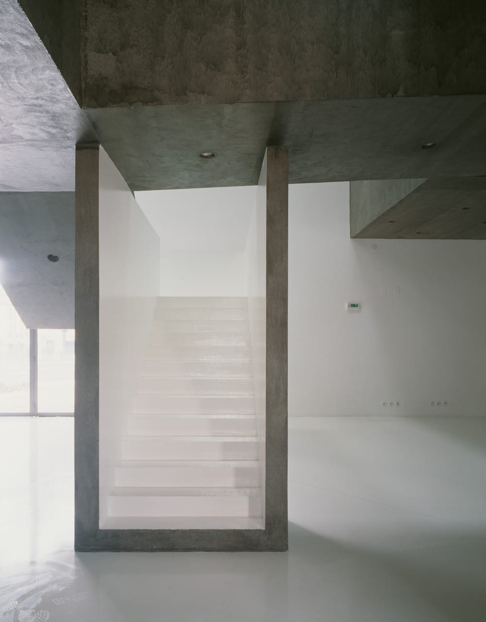 White staircase framed by a dark concrete ceiling, column and cantilevered volume inside Casa dos Cubos.