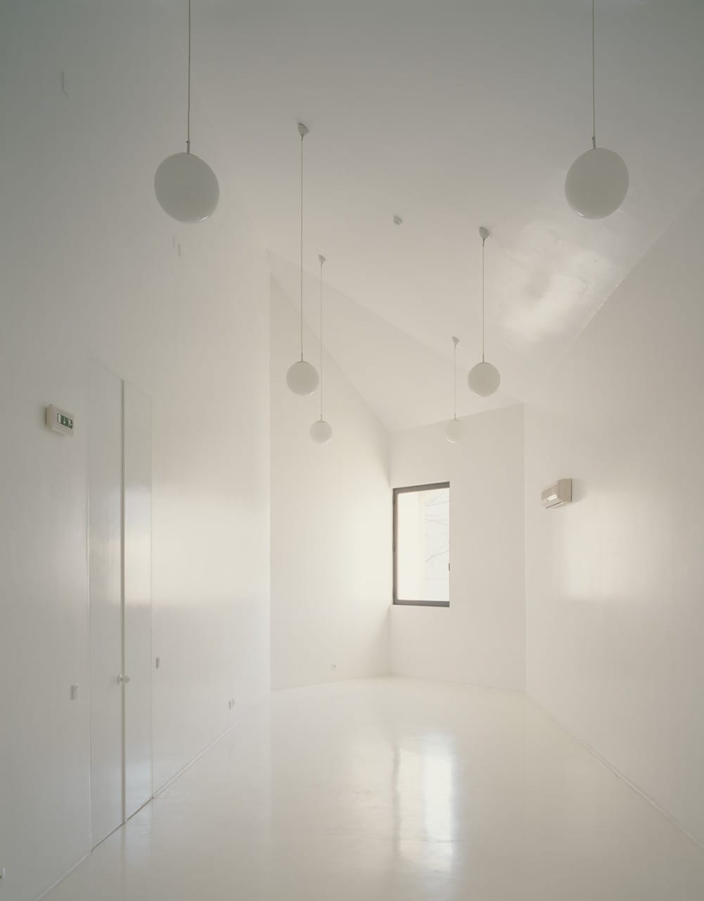 Bright white Casa dos Cubos room with spherical pendant lights and a tall narrow window.