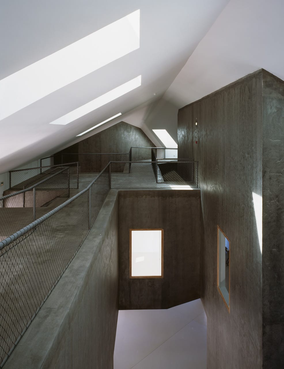 Mesh-railed walkway threading between dark concrete room volumes beneath skylights at Casa dos Cubos.