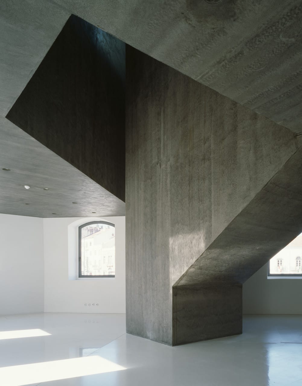 Dark concrete cantilever and staircase rising above the glossy white floor at Casa dos Cubos.