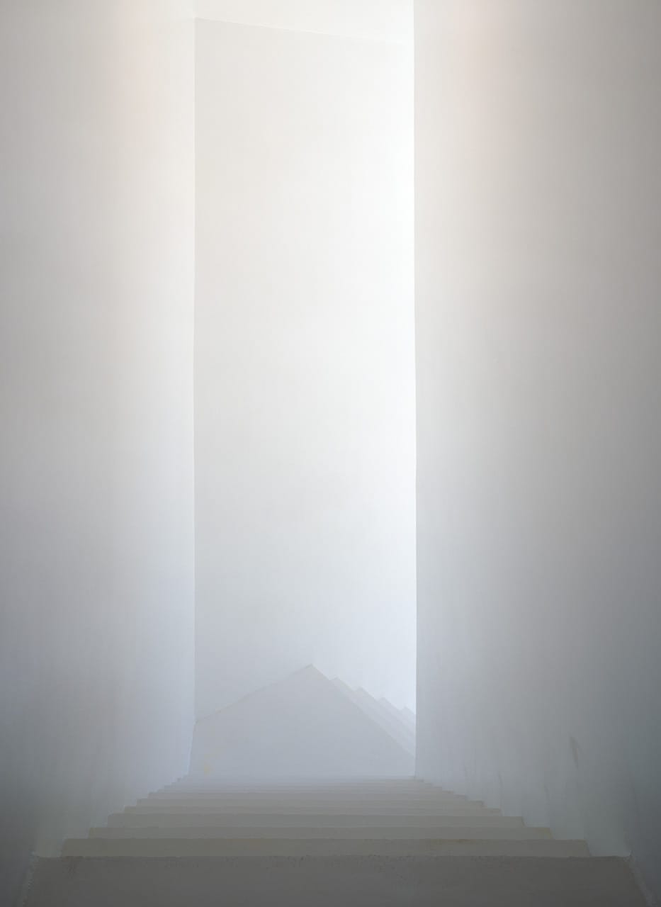 White stairwell descending through a tall narrow opening at Casa dos Cubos.