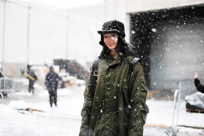 Wide portrait of Xiao Wen Ju wearing an olive parka and black cap on a snowy New York street.