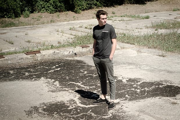 Model wearing a charcoal Deadwood T-shirt and cropped grey trousers in an industrial lot.