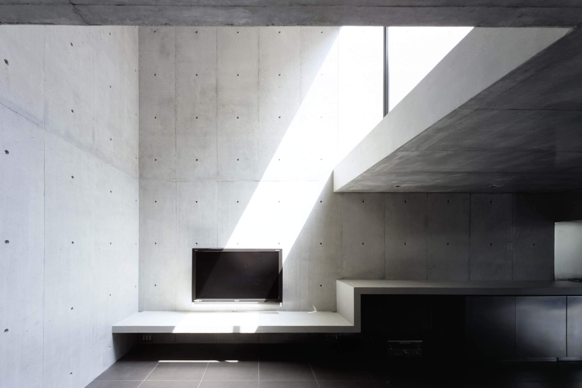 Double-height living room in 2COURTS HOUSE with a diagonal shaft of daylight across exposed concrete.