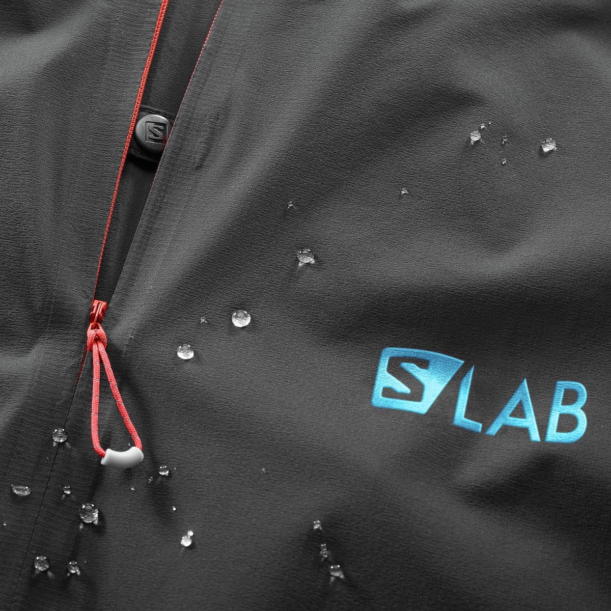 Close-up of the black Salomon S/LAB shell with water beading on the face fabric, turquoise S/LAB chest mark, and red zipper pull. SAL00V8 is filename-supported; exact model remains unconf...