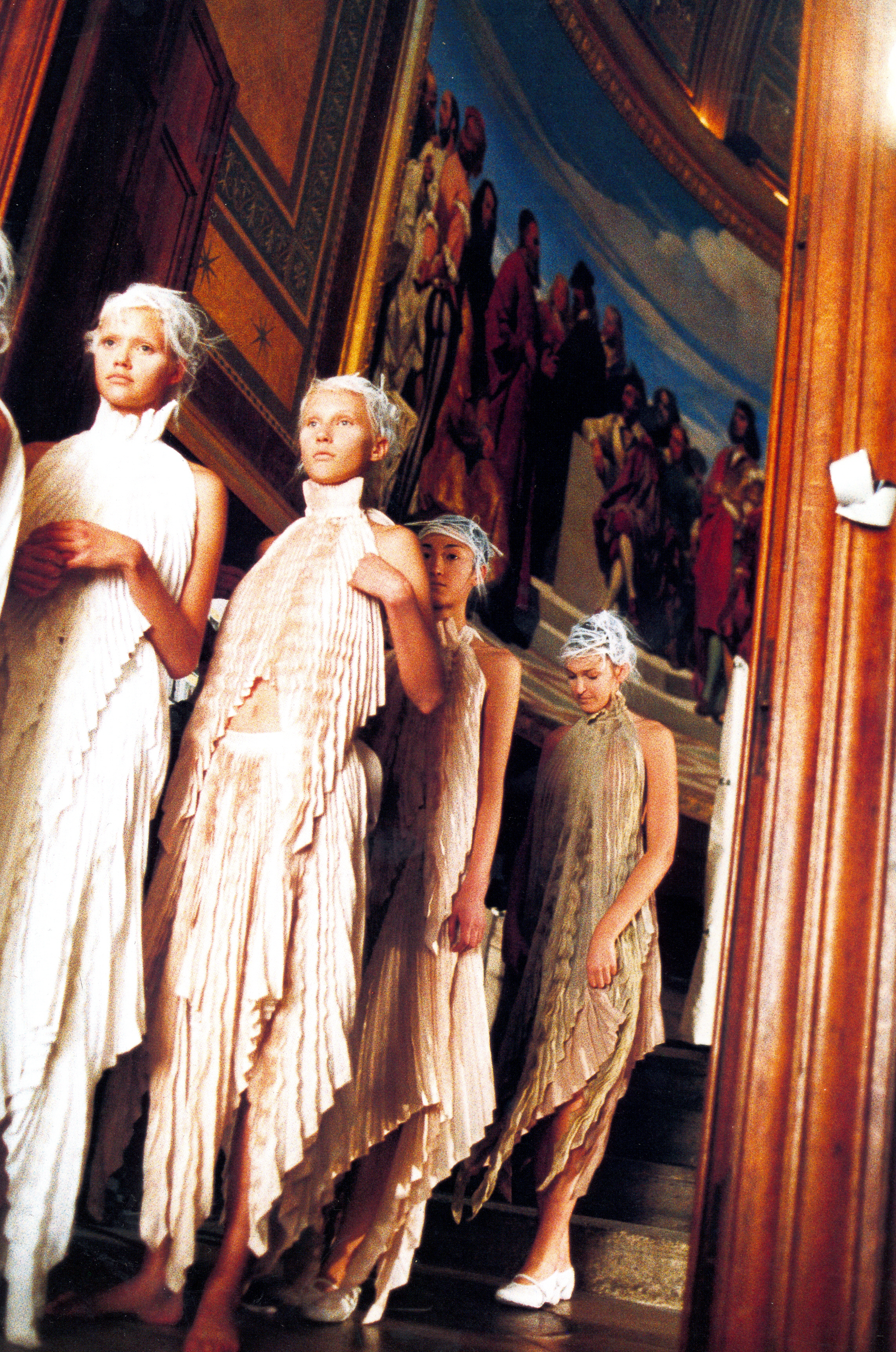Models in pale pleated dresses descending an ornate staircase.