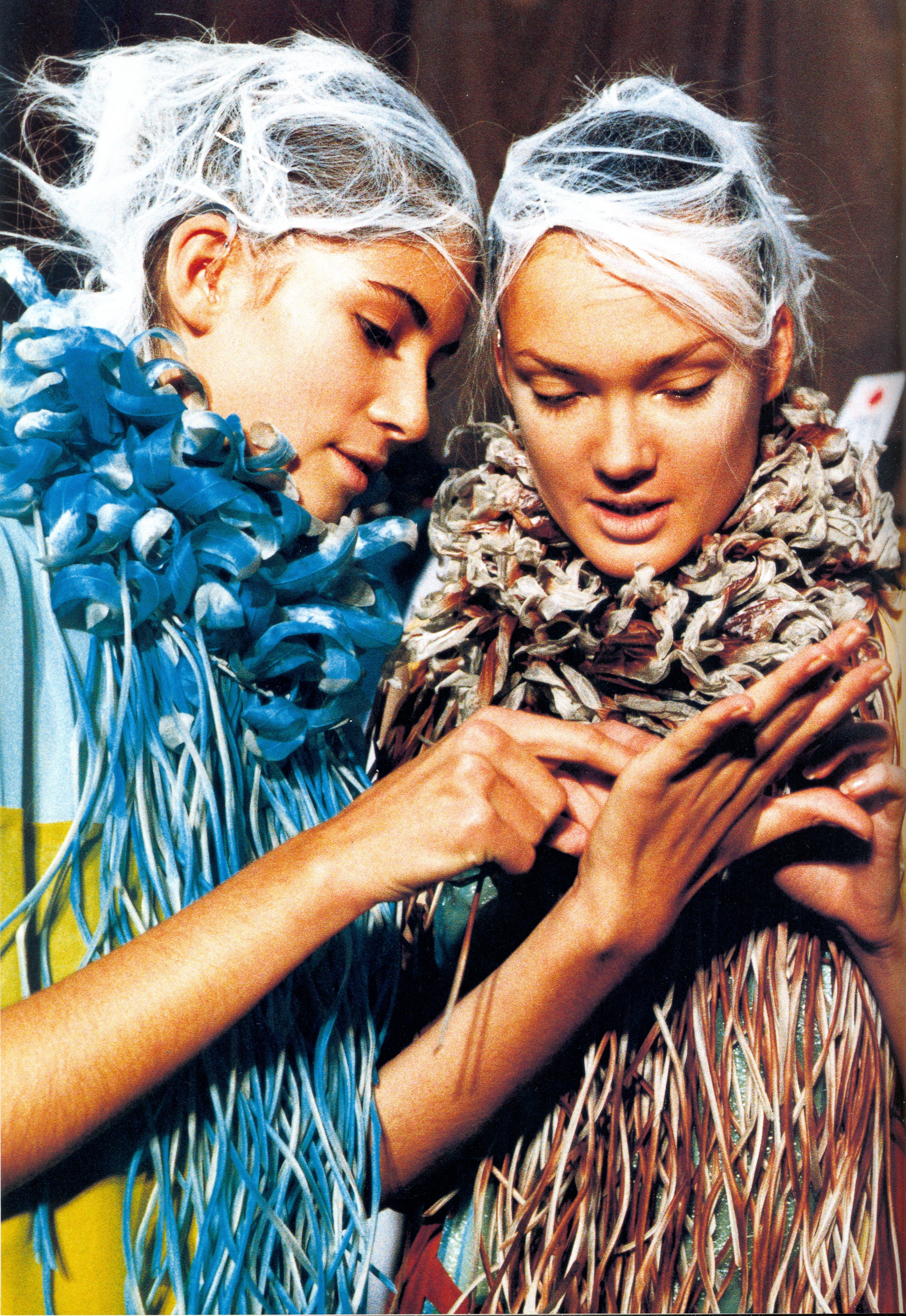 Two women wearing white fibrous hair styling and densely fringed blue and brown clothing.