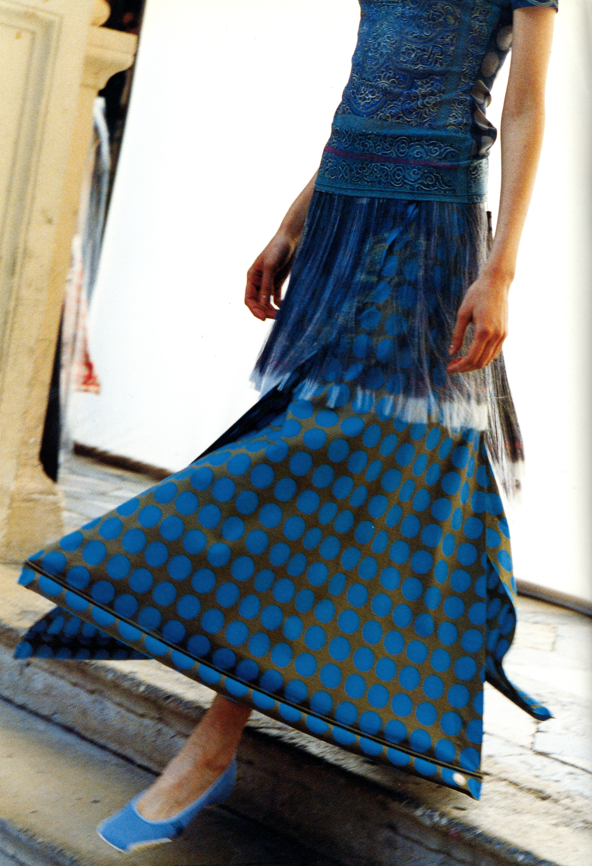 Cropped walking figure in a blue patterned top, sheer overskirt, and blue polka-dot skirt.