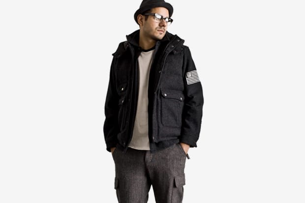 Model in a charcoal Stussy Japan hooded field jacket with a black-and-white flag patch on the sleeve.