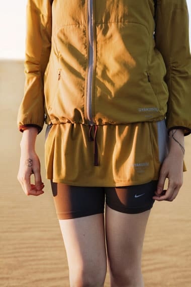 Woman in a mustard Gyakusou SS2012 running top and shorts against pale sand dunes