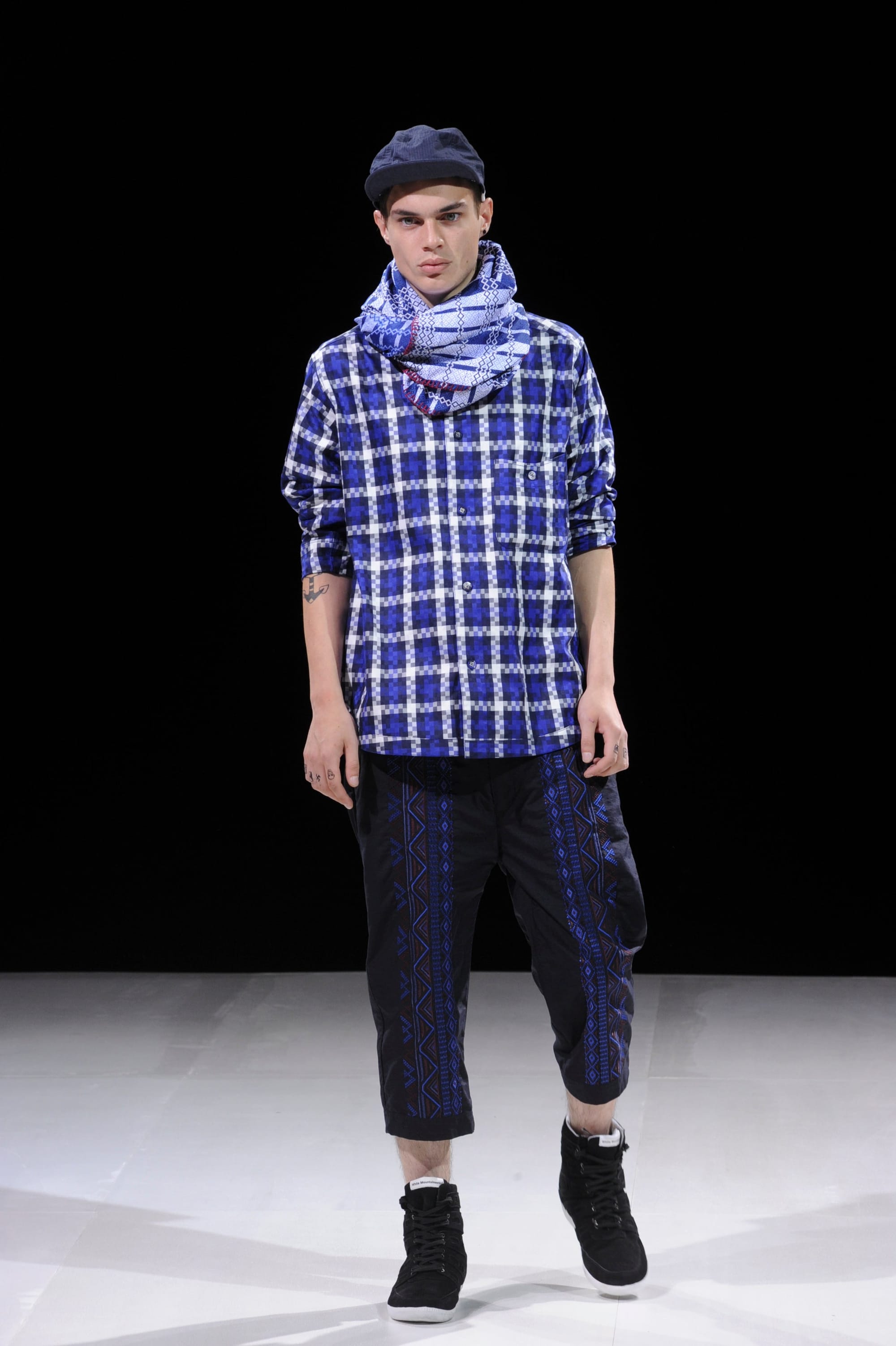 Model in a blue plaid scarf and tunic with black cropped trousers and black boots.