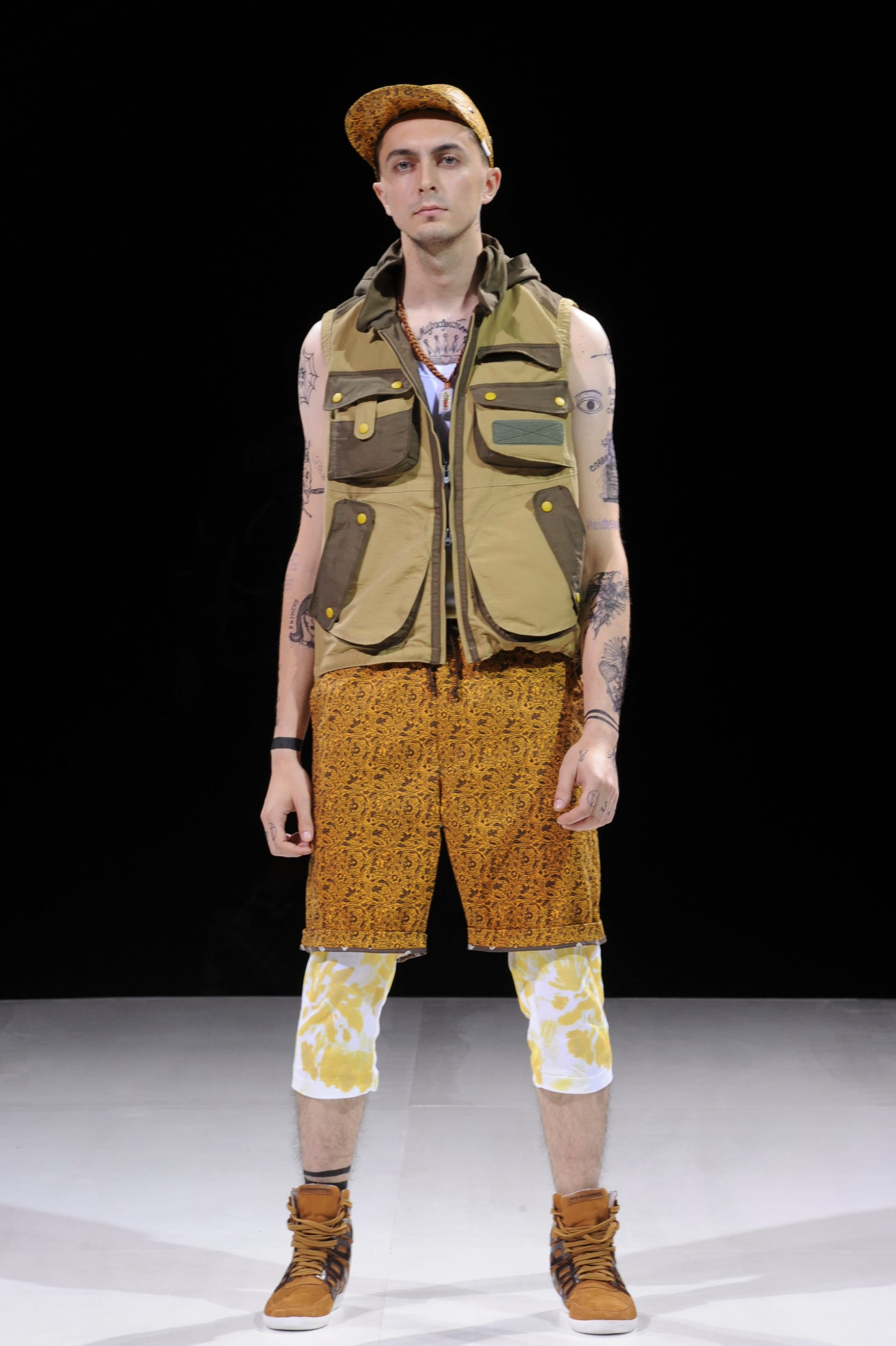 Model in an olive utility vest and mustard patterned shorts layered over white printed leggings.