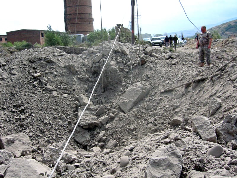 Large bomb crater amid rubble and broken cables in Gori, with soldiers and vehicles nearby.
