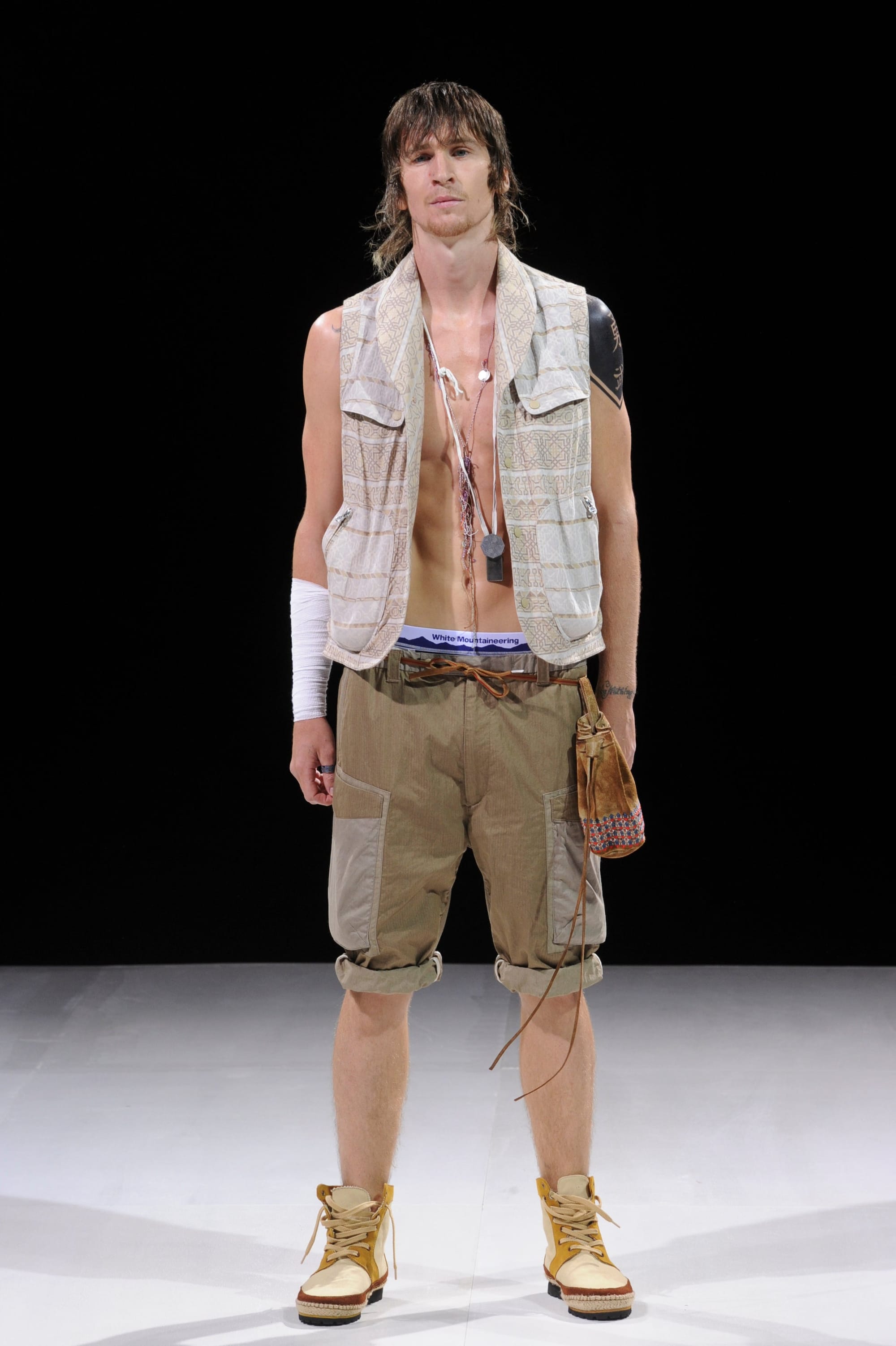 Model in a pale plaid utility vest and tan cargo shorts with a white arm wrap.