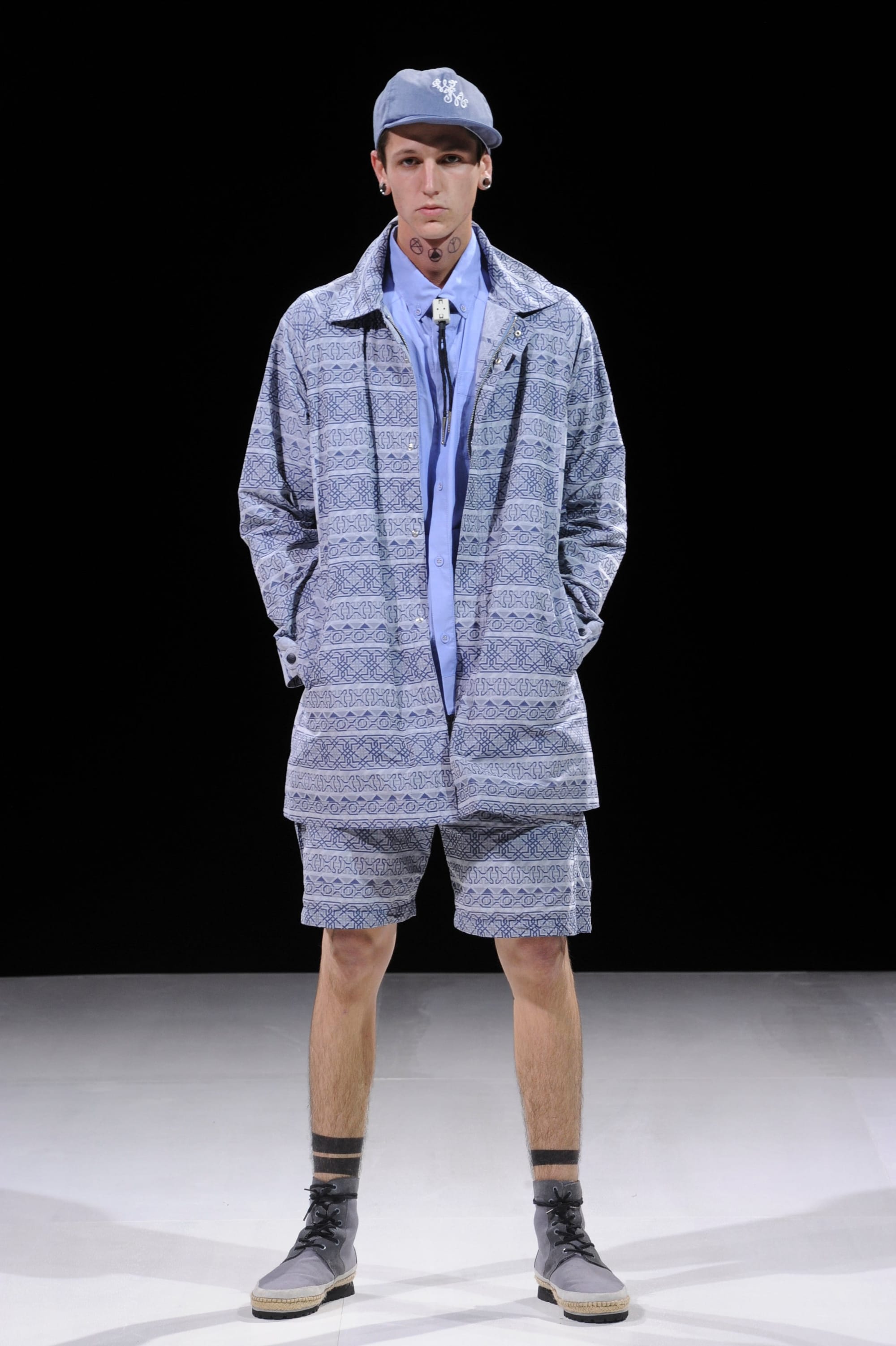 Model in a blue-grey patterned coat and matching shorts with a navy cap.