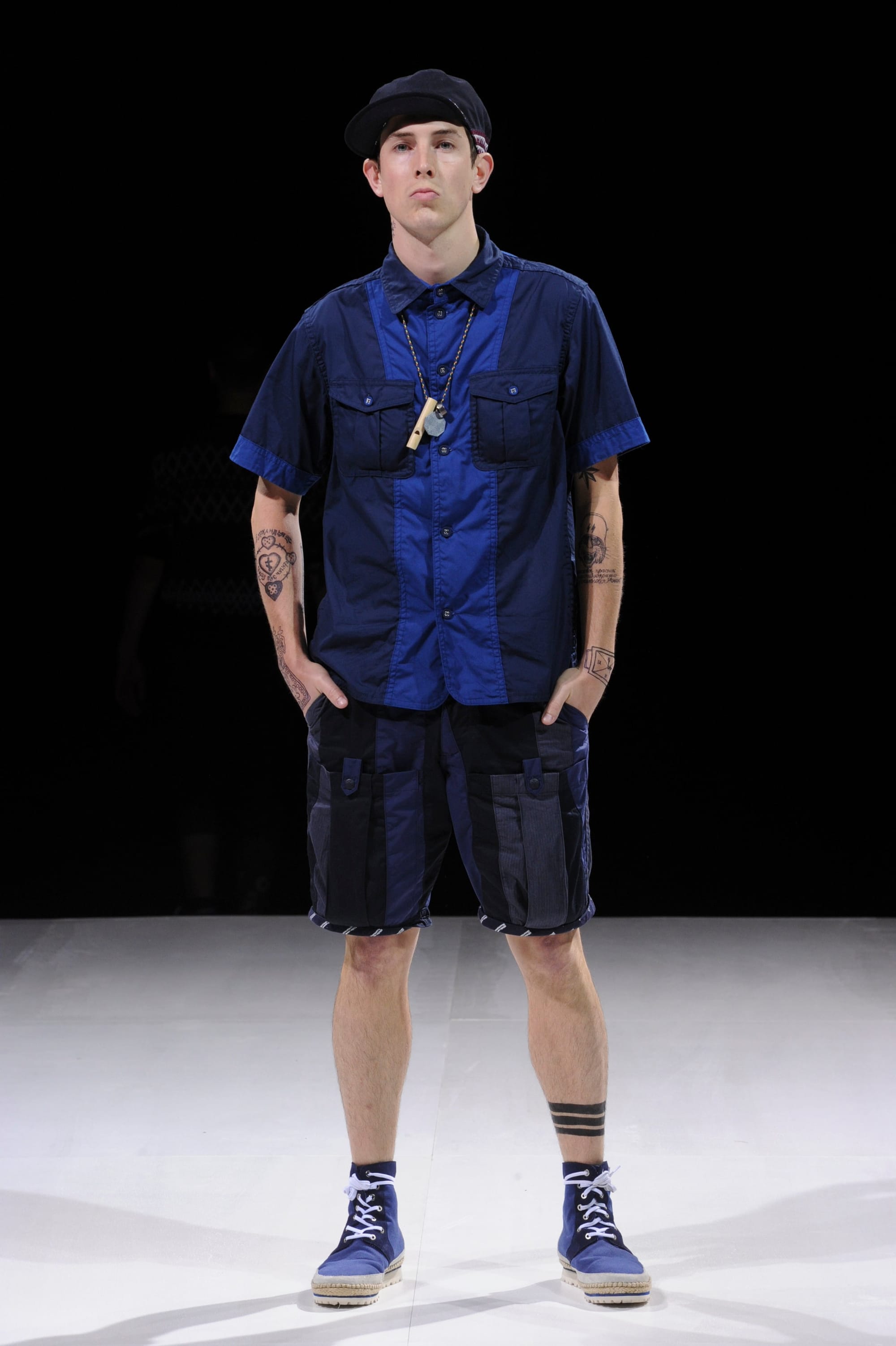 Model in a blue utility shirt and dark cargo shorts with a black beret.