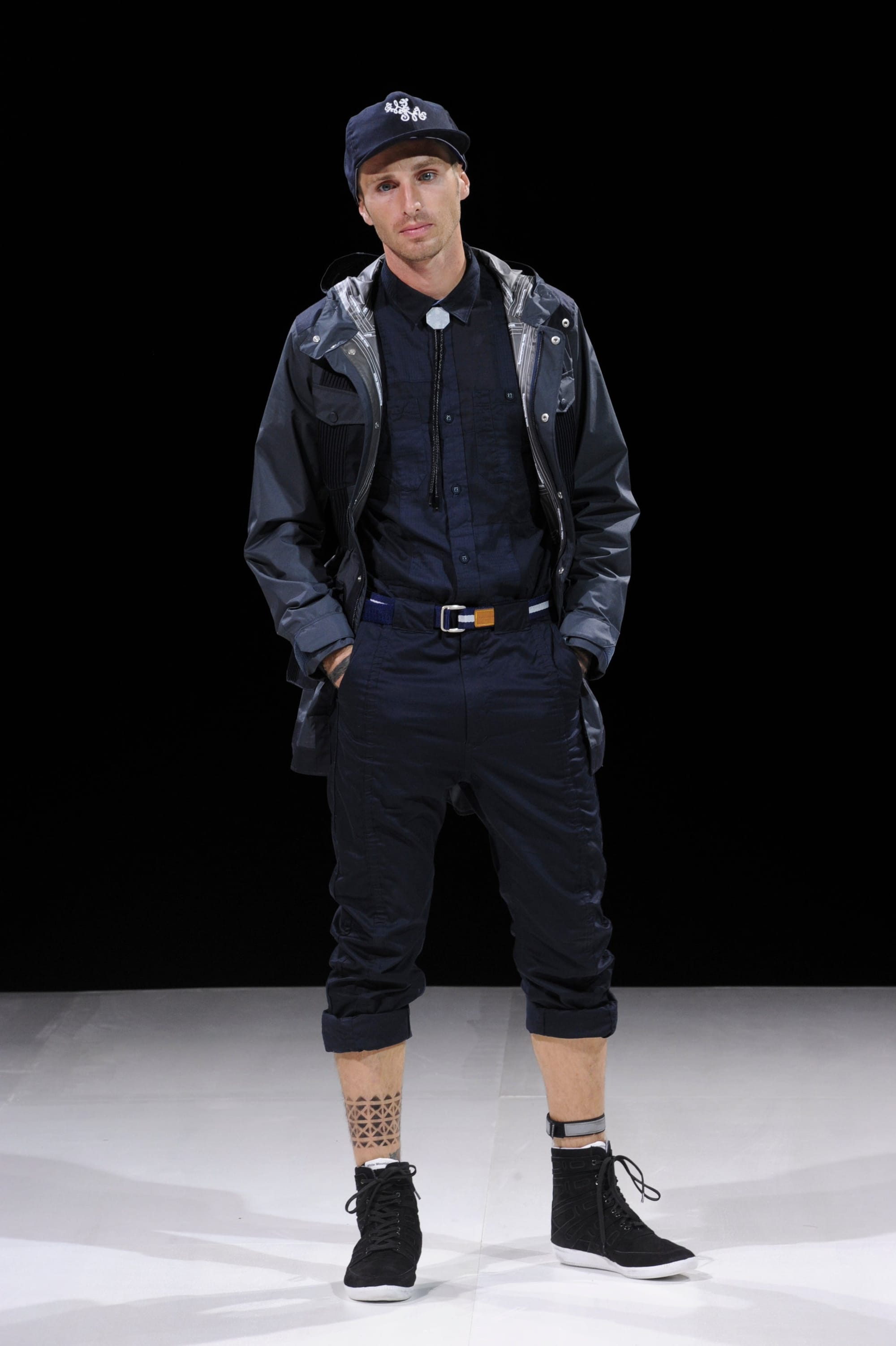 Model in a black technical jacket and black cropped trousers with a navy cap.