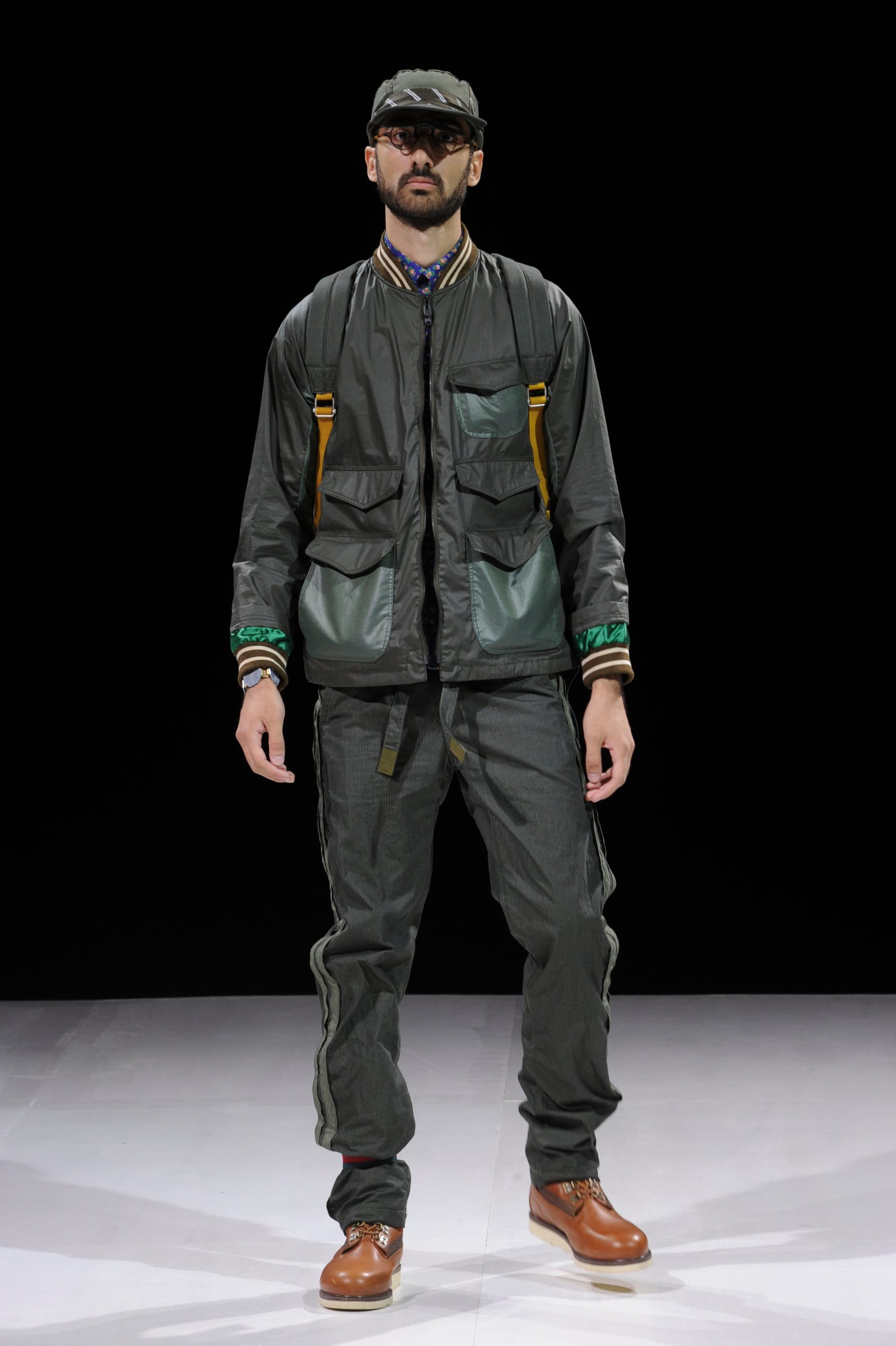 Model in a dark olive technical jacket and trousers with green pockets, yellow straps, cap, and sunglasses.