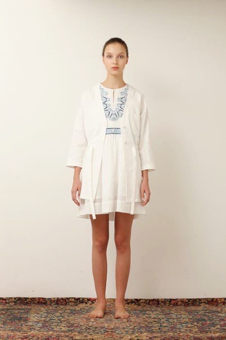 White Mountaineering womenswear model in a white open coat over a white dress with blue embroidery.