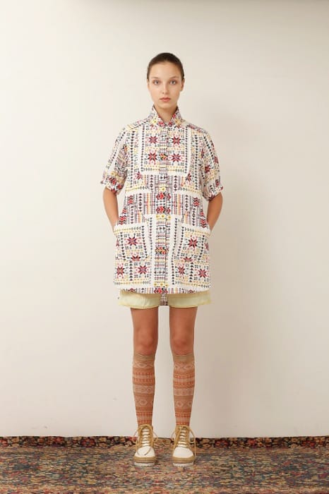 White Mountaineering womenswear model in a multicolour patterned coat, cream shorts, patterned knee socks and tan high-top sneakers.