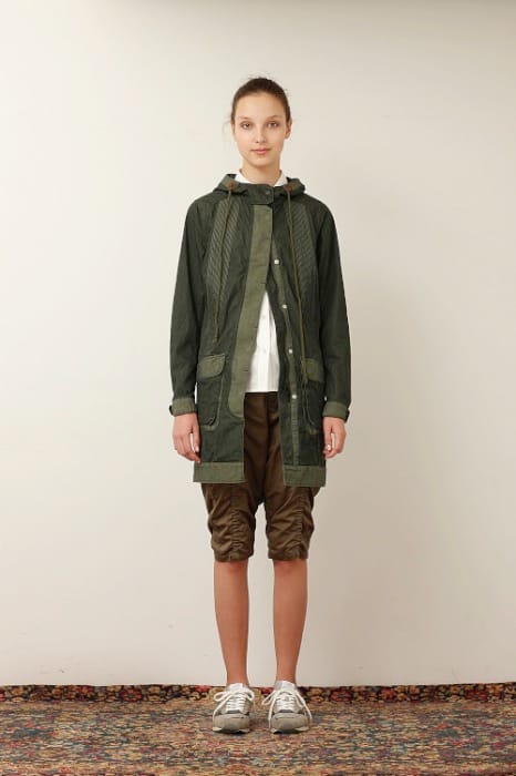 White Mountaineering womenswear model in an olive hooded parka, white shirt and cropped brown trousers.