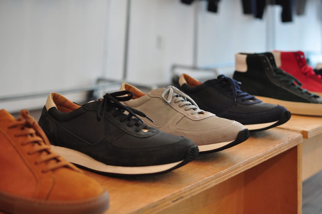 Black, gray and navy Common Projects running sneakers displayed with leather high-tops on wooden showroom tables.