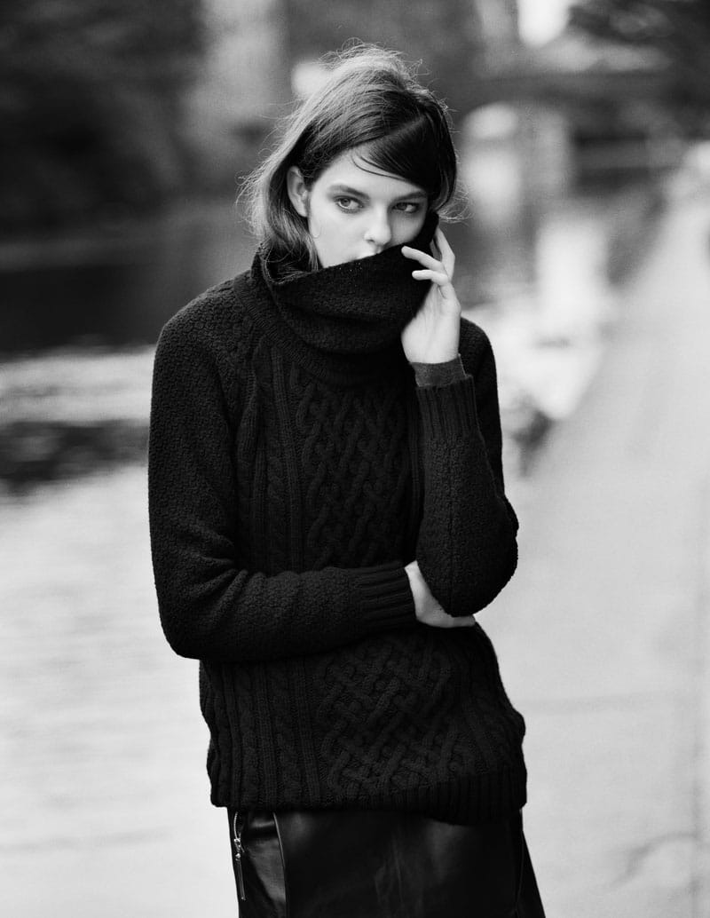 Kia Low raising the collar of a black cable-knit John Smedley jumper in a black-and-white canal-side portrait.